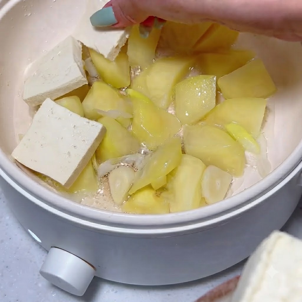 Rodajas de tofu ordenadas cuidadosamente sobre una capa de trozos de patata cocida y ajo dentro de una olla blanca.