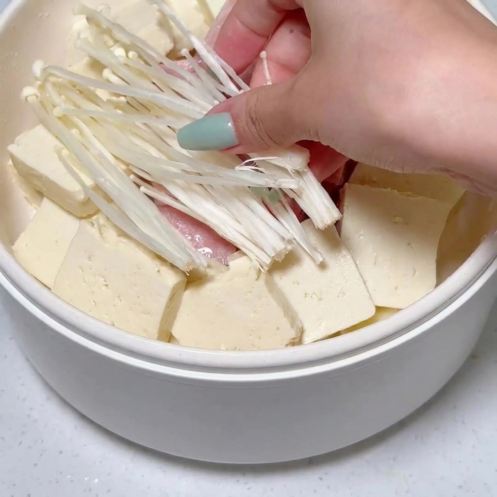 Champiñones enoki siendo colocados en la olla entre el tofu y las rodajas de carne enlatada.