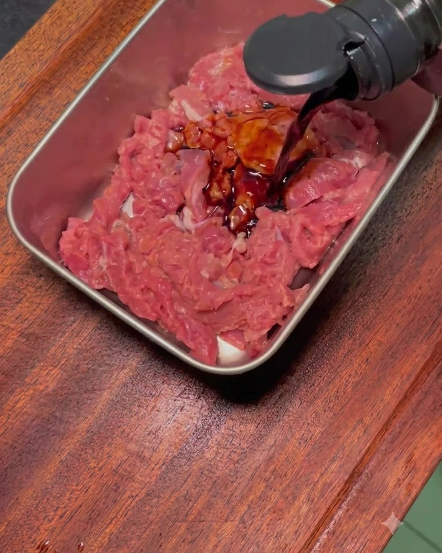 Salsa oscura de marinado vertiéndose sobre ternera cruda laminada en una bandeja metálica.
