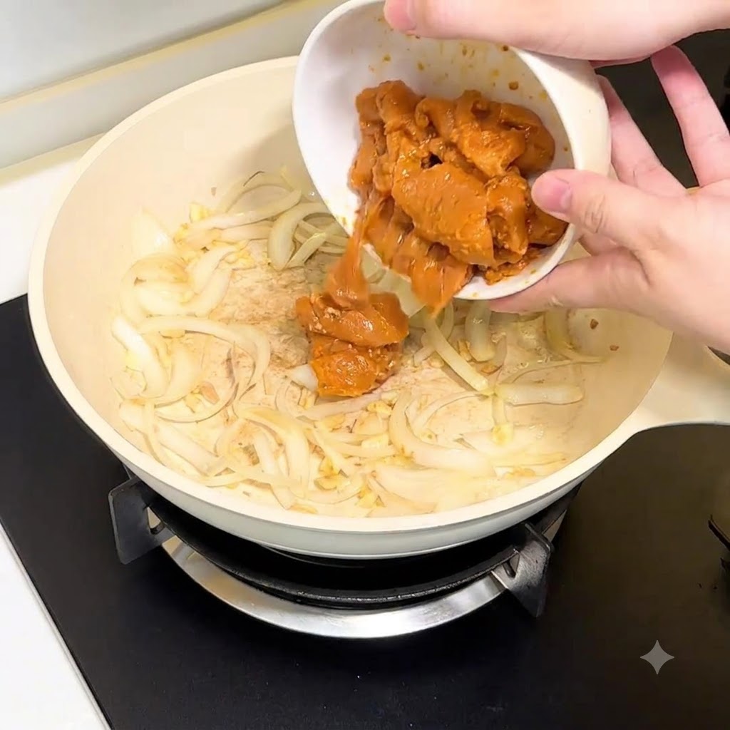 Trozos de pechuga de pollo siendo vertidos desde un bol blanco hacia la sartén con cebolla y ajo.