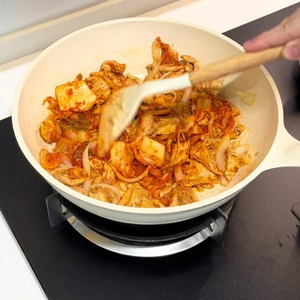 Kimchi y pollo salteándose juntos en una sartén blanca mediante una espátula.