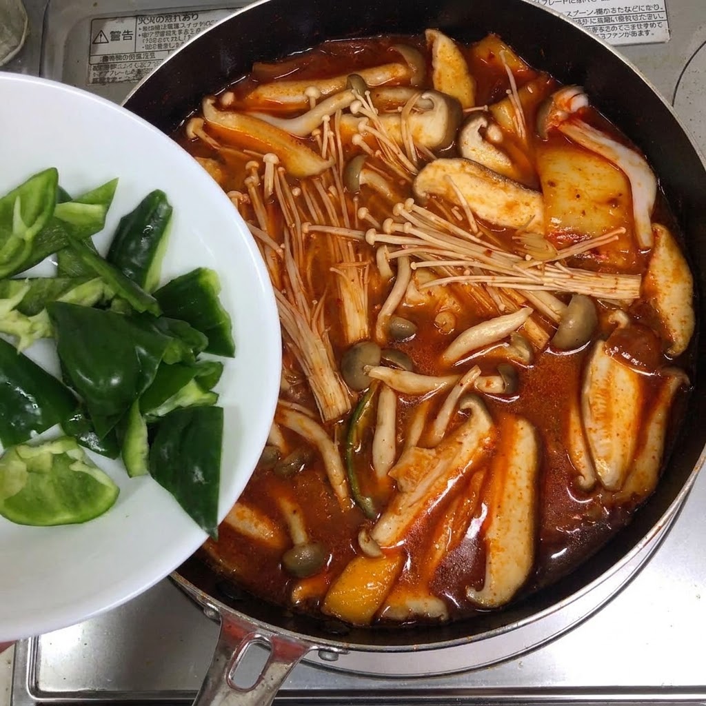 Rodajas de pimientos verdes siendo añadidas desde un plato blanco a una olla de sopa roja de kimchi.