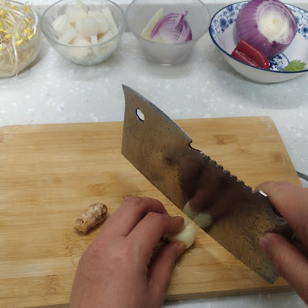 Dientes de ajo y raíz de jengibre fresco cortados en una tabla de cortar al lado de tazones con ingredientes.