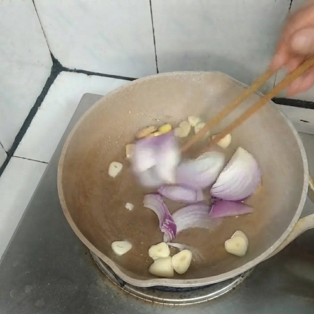 Cebollas rojas en rodajas, ajo y jengibre salteándose juntos en una sartén con palillos de madera.
