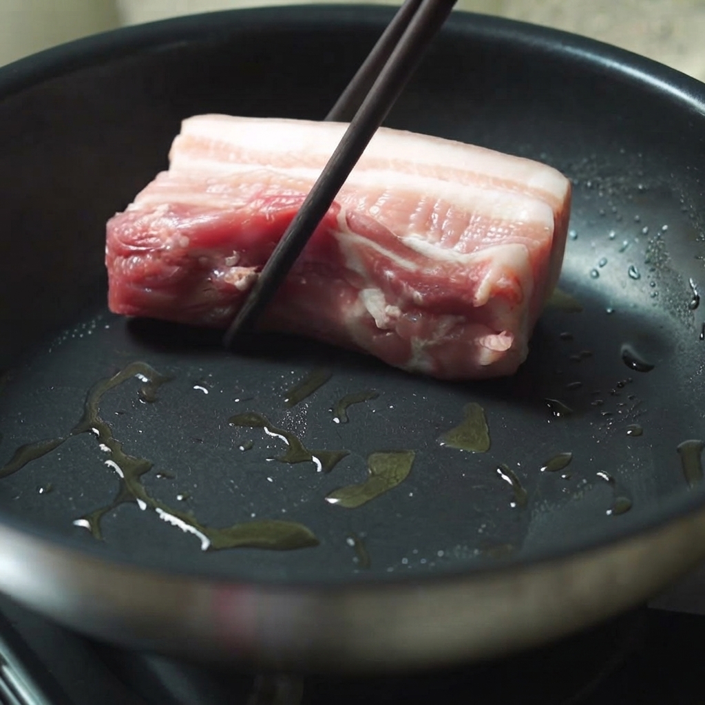 Un trozo grueso de panceta de cerdo siendo colocado en una sartén caliente con aceite usando palillos.