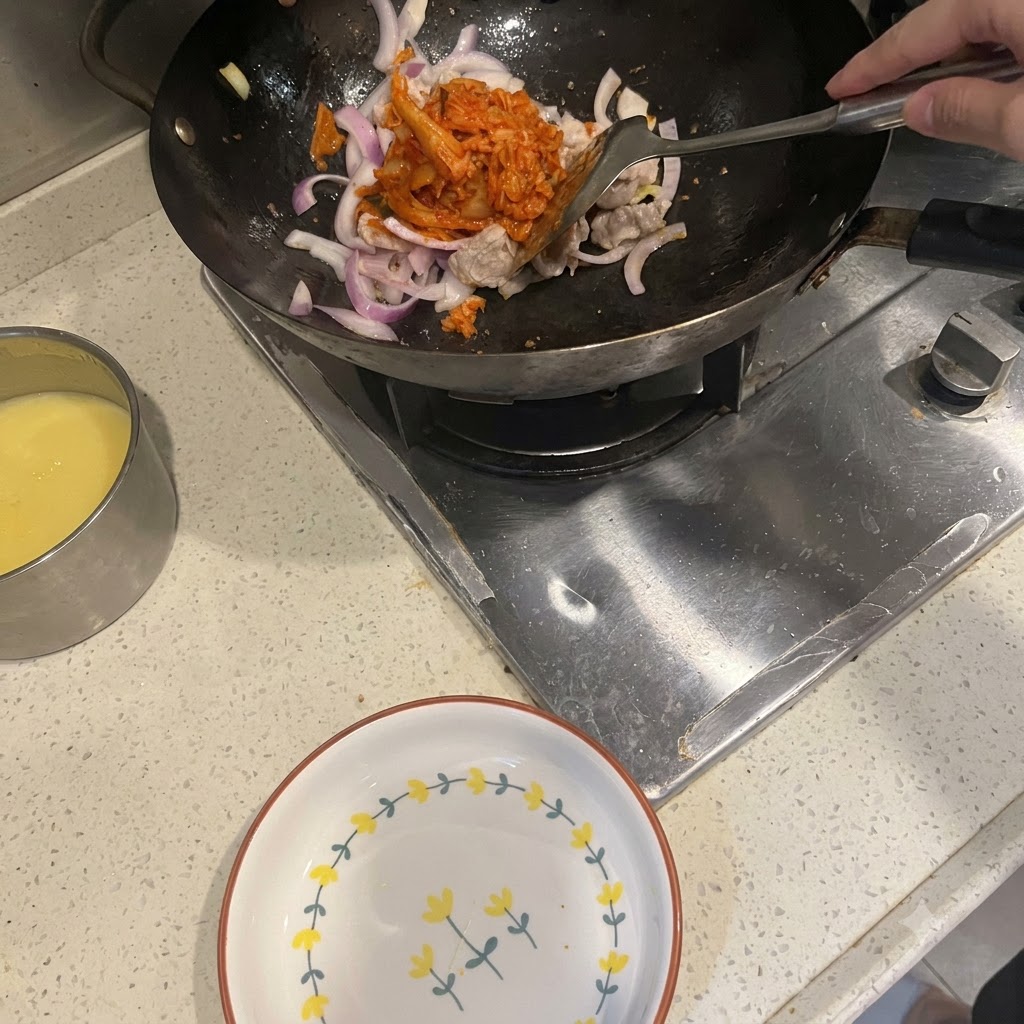 Una espátula añadiendo kimchi rojo brillante en un wok que contiene panceta de cerdo salteada y cebollas moradas.