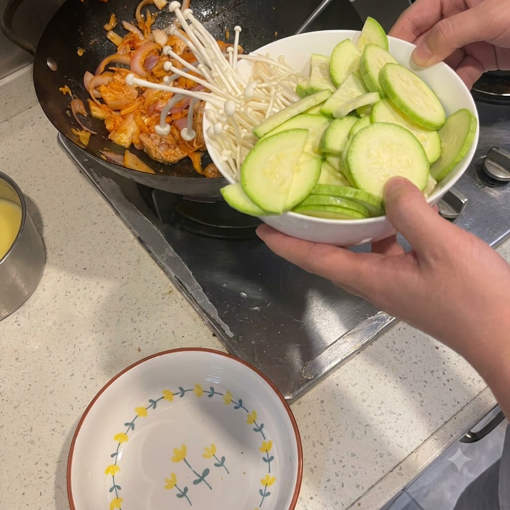 Calabacín cortado y setas enoki siendo vertidos desde un bol hacia un wok que contiene cerdo y cebollas salteadas.