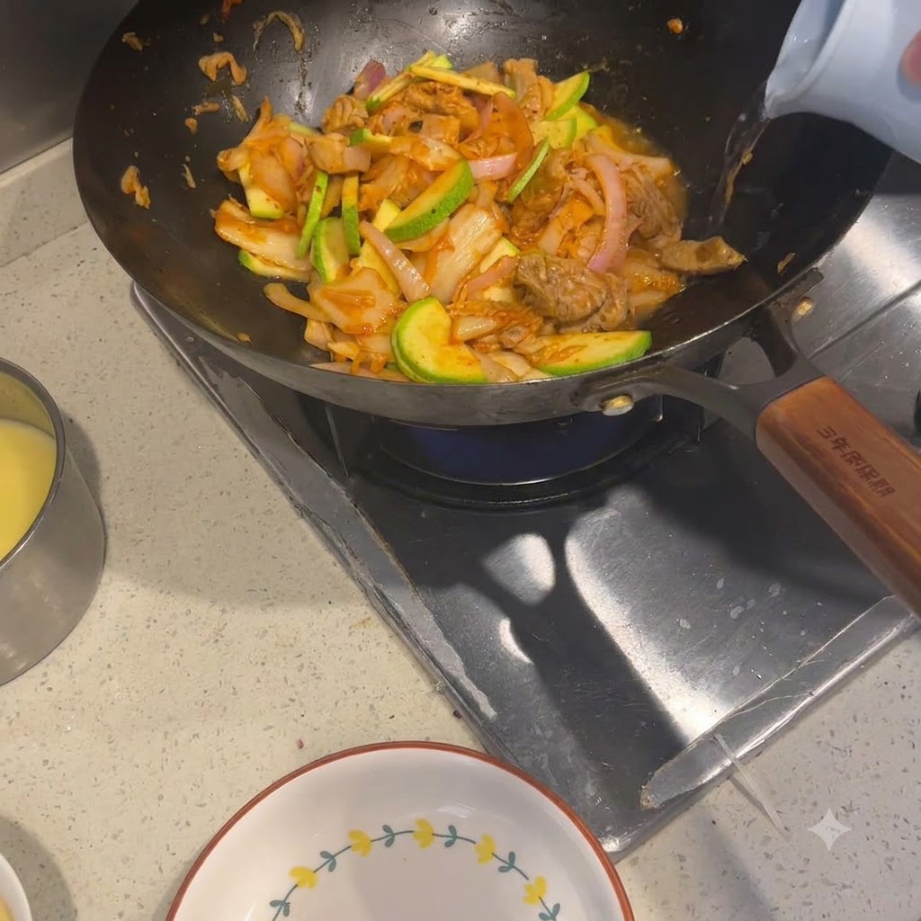 Agua caliente siendo vertida desde un hervidor de tonos claros en un wok lleno de ingredientes salteados.
