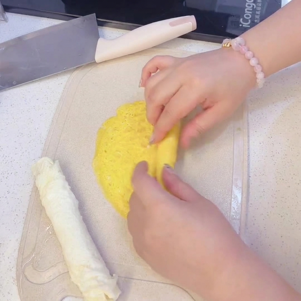Manos enrollando una fina crepa de huevo amarillo sobre una tabla de cortar, junto a una crepa de huevo blanco ya enrollada.