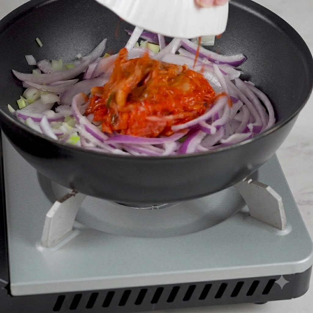 Kimchi coreano siendo añadido desde un cuenco a la sartén que contiene las cebollas salteadas.