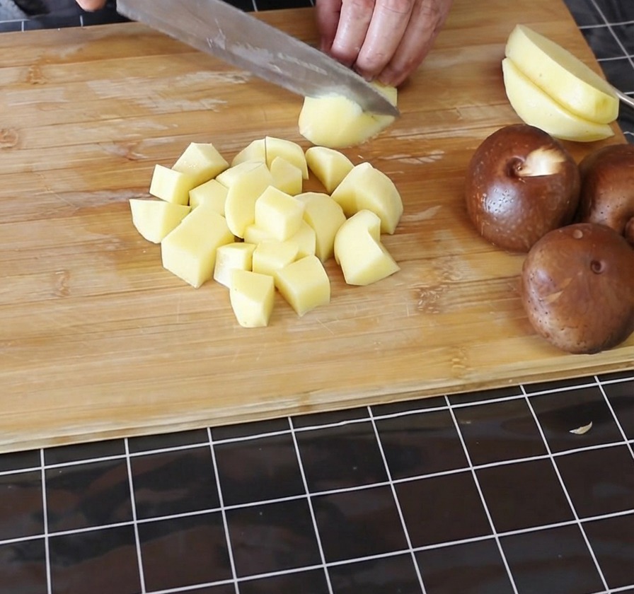 Manos cortando patatas amarillas peladas en cubos pequeños sobre una tabla de cortar de bambú.