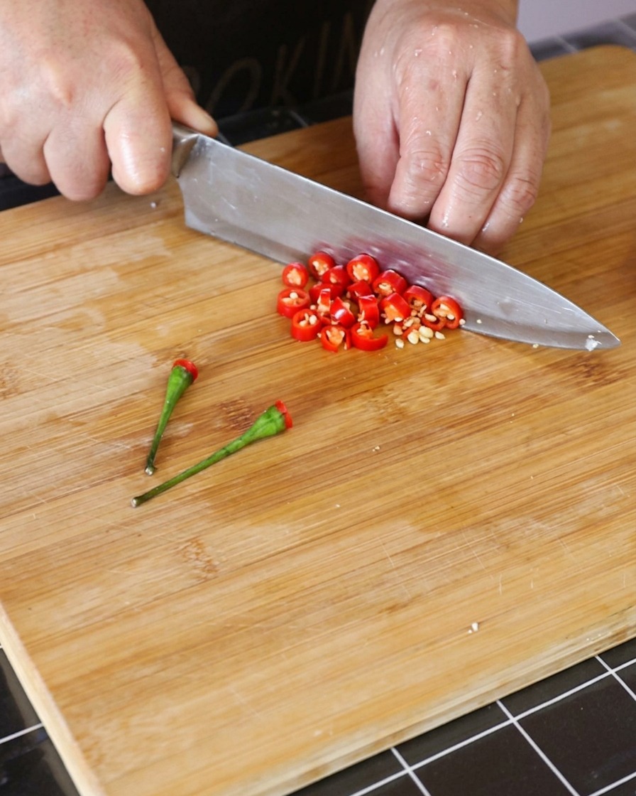 Chiles tailandeses rojos frescos siendo cortados en pequeños aros sobre una tabla de cortar de madera.