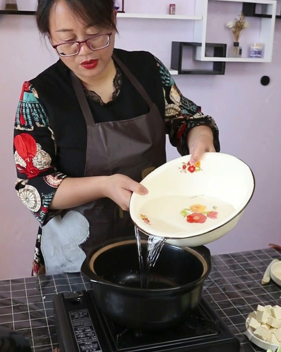 Agua clara siendo vertida desde un cuenco decorativo en una olla de cerámica negra sobre una estufa portátil.