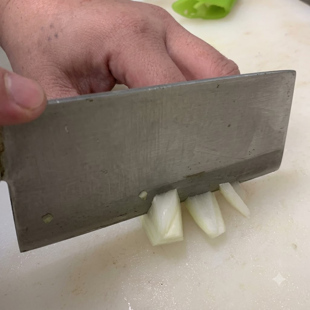 Un cuchillo cortando una cebolla en trozos medianos sobre una tabla de cortar blanca.