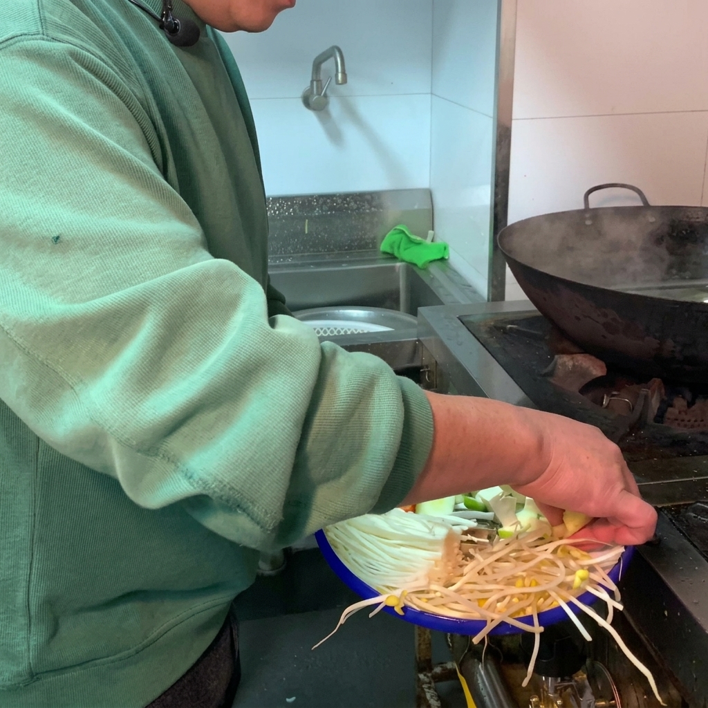 Una variedad de verduras frescas y tofu blanco siendo agregados a una olla de barro oscura llena de agua.