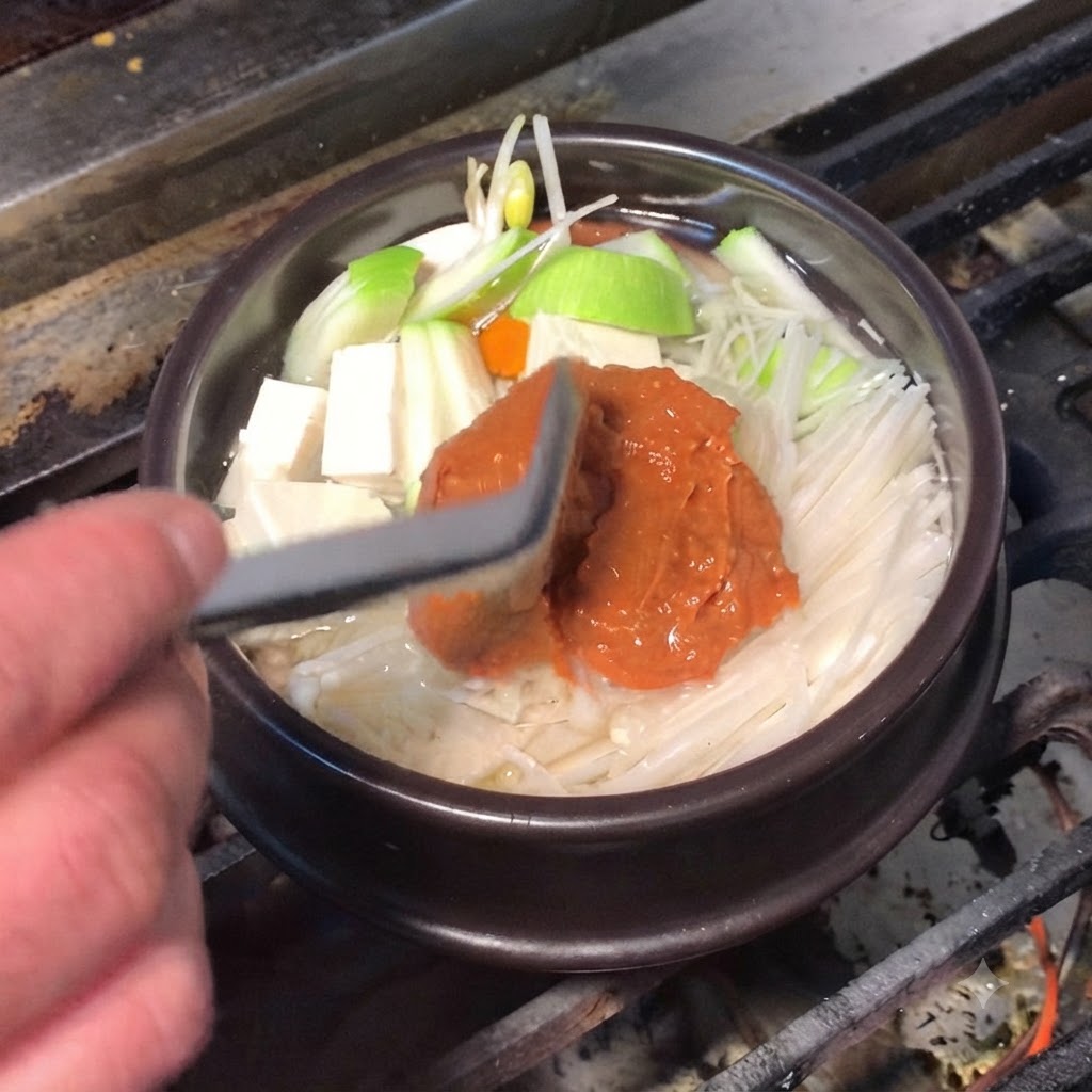 Una espátula de silicona agregando rica pasta de soja marrón y pasta de ají roja en la olla de cocción.