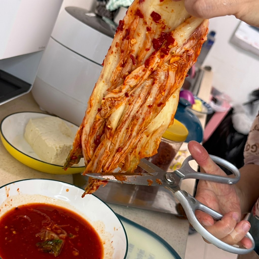 Un par de tijeras de cocina plateadas cortando un trozo de kimchi rojo sobre un plato blanco lleno de salsa roja picante.