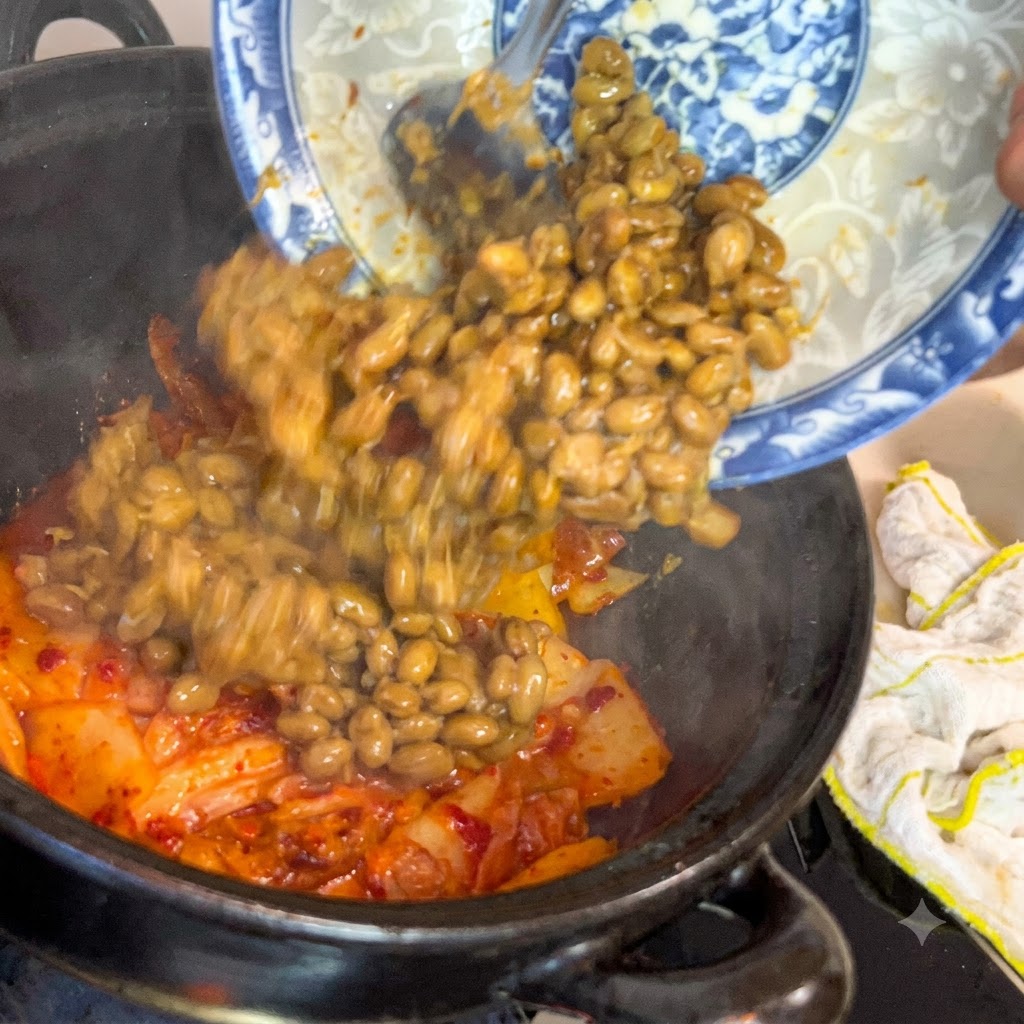 Soja fermentada deslizándose desde un plato decorativo azul y blanco hacia una olla de cocina oscura llena de kimchi chisporroteante.