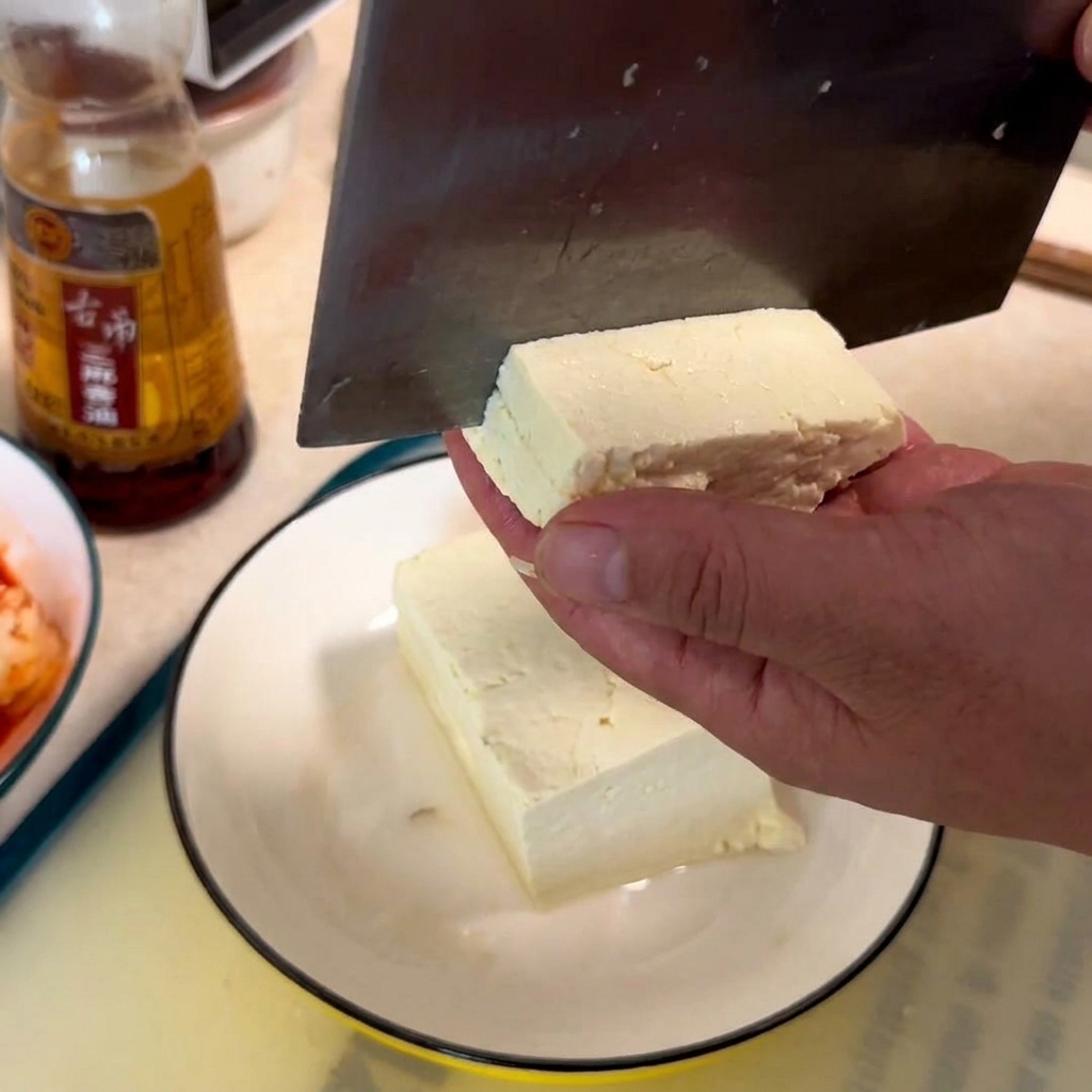 Una mano sosteniendo un bloque de tofu blanco mientras un cuchillo de carnicero lo corta en cubos sobre un plato blanco.