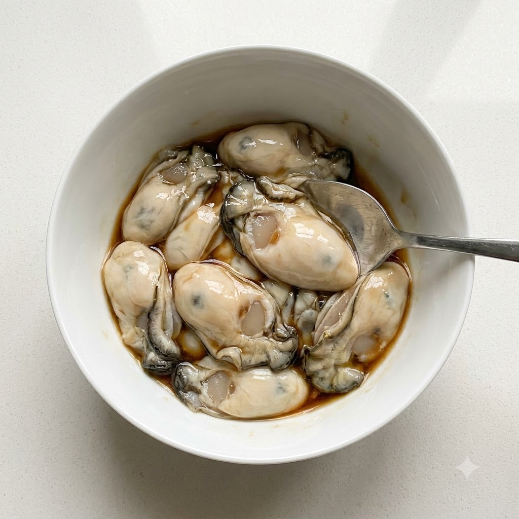 Ostras limpias en un tazón blanco siendo sazonadas con salsa de pescado y pimienta blanca en polvo usando una cuchara.
