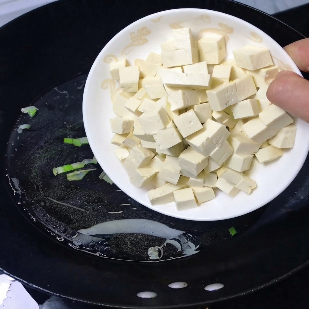 Un plato de tofu blanco en cubos siendo vertido en un wok negro con agua hirviendo.