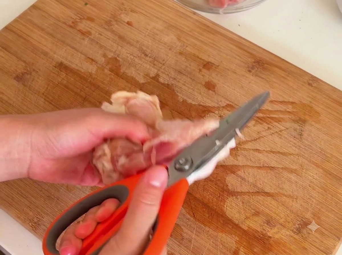 Unas tijeras de cocina de color naranja cortando la carne de un hueso de muslo de pollo crudo sobre una tabla de madera.