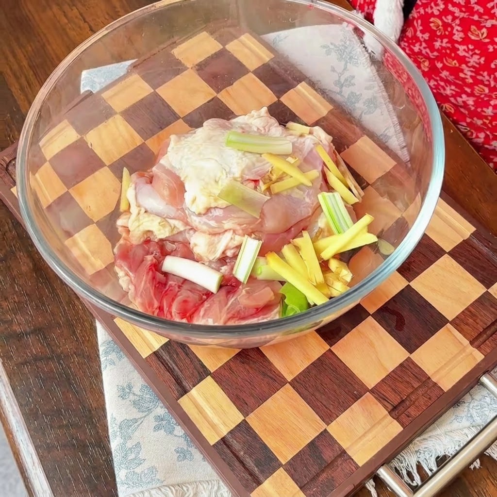 Trozos de pollo crudo en un bol de cristal cubiertos con cebolla roja en rodajas, cebolletas y tiras de jengibre.
