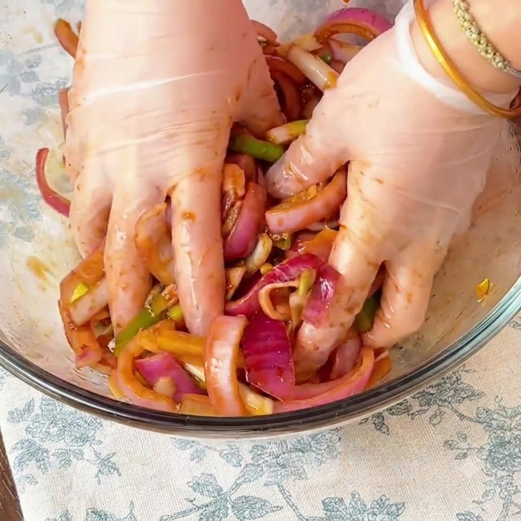 Manos con guantes masajeando a fondo la marinada y la cebolla roja en rodajas en el pollo dentro de un bol de cristal.