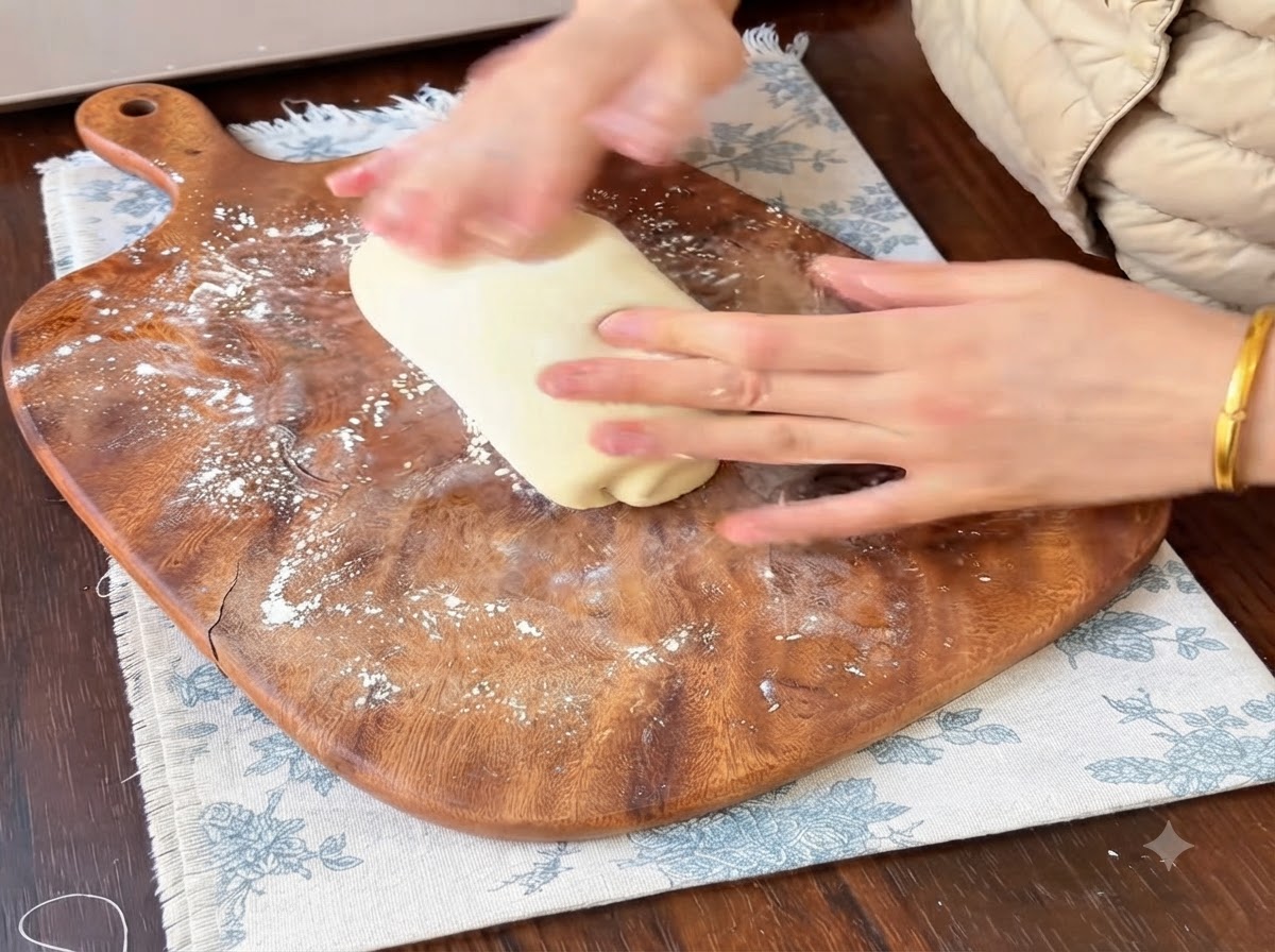 Manos enrollando y dando forma a una porción de masa de pan suave sobre una tabla de cortar de madera espolvoreada con harina.