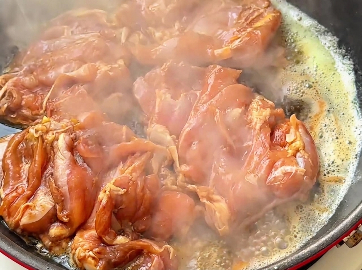 Trozos de contramuslo de pollo crudo marinado friéndose en una sartén caliente con mantequilla y vapor saliendo.