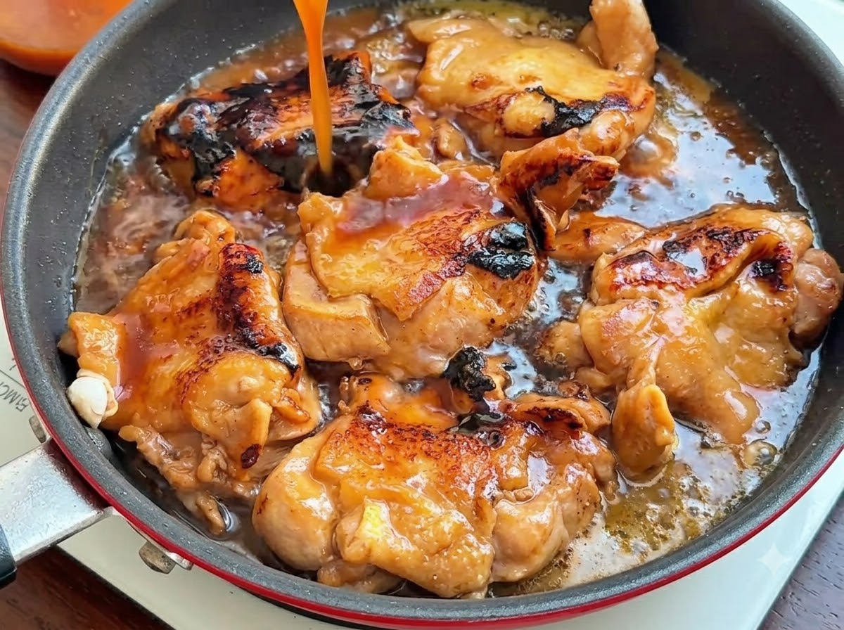 Un chorrito de salsa de color marrón oscuro vertiéndose desde arriba en una sartén llena de contramuslos de pollo dorados.