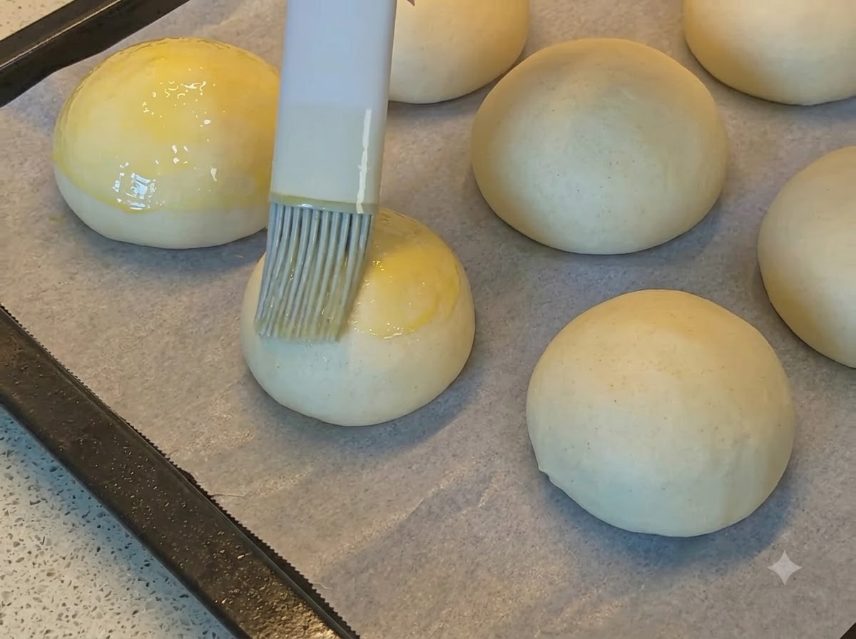 Un pincel de repostería aplicando huevo batido dorado sobre bolas de masa de pan redondas y lisas en papel de hornear.