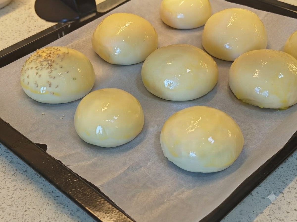 Semillas de sésamo siendo espolvoreadas sobre una bandeja de panecillos de pan brillantes pintados con huevo.