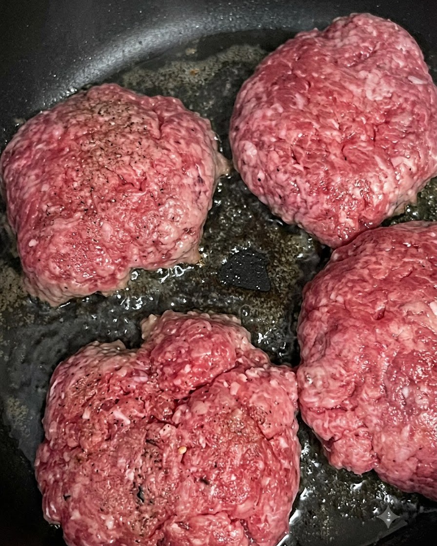 Cuatro hamburguesas de ternera crudas cociéndose y dorándose en una sartén caliente.