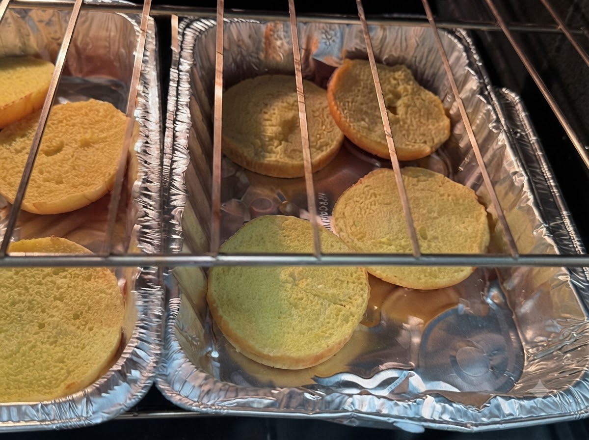 Mitades de pan de hamburguesa reposando en bandejas de papel de aluminio dentro de las resistencias del horno.