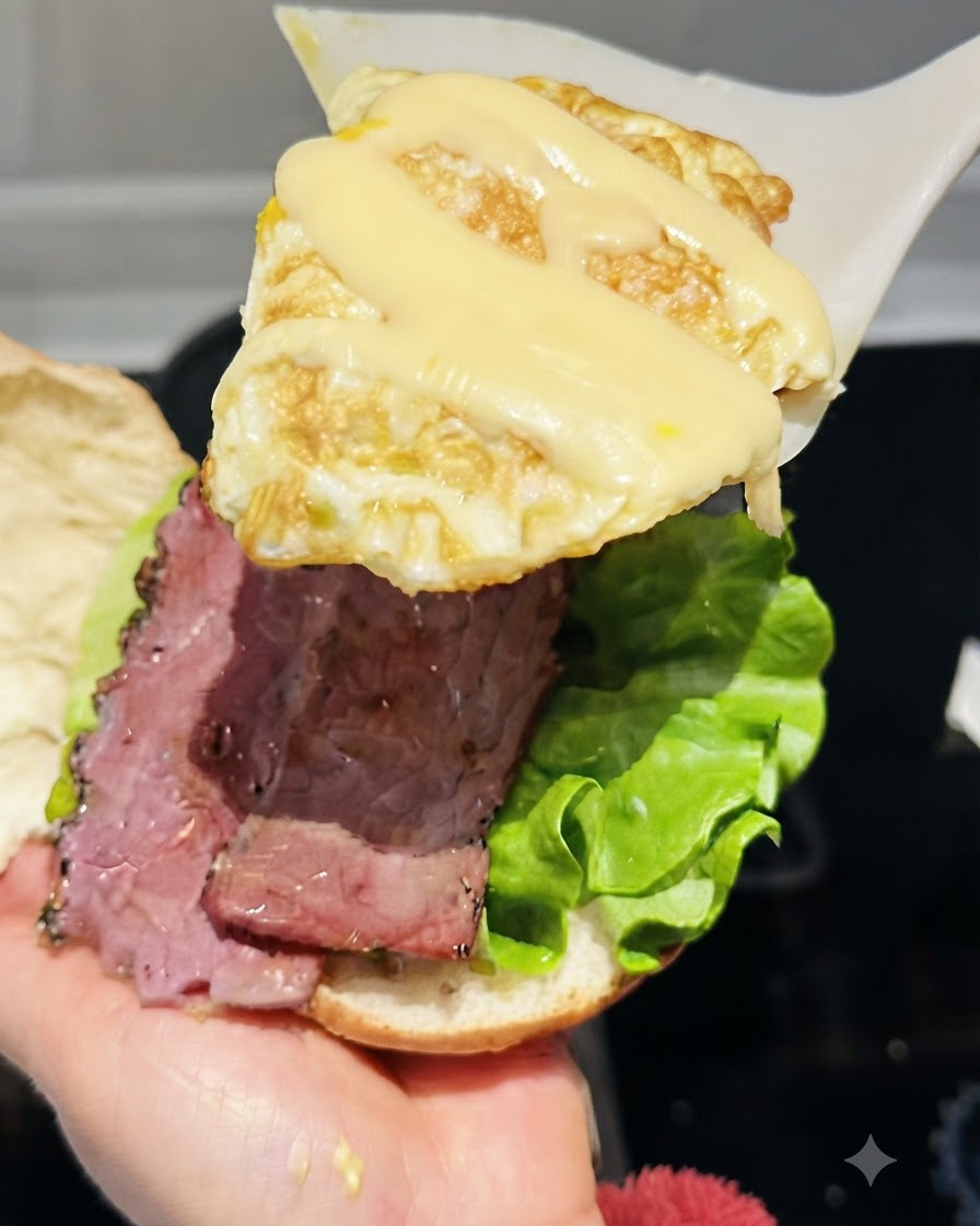 Una espátula levantando un huevo frito cubierto con queso en tiras fundido sobre el armado de la hamburguesa.