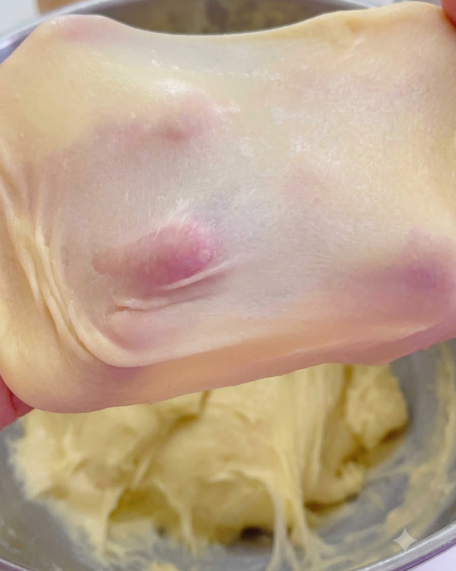 Masa de brioche estirada que muestra una membrana de tipo ventana delgada, translúcida y sostenida por los dedos.