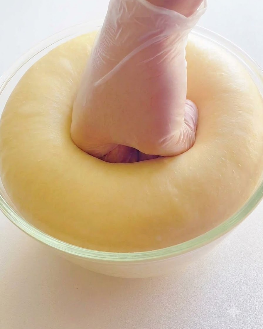 Una mano con un guante transparente presionando el centro de una masa esponjosa y bien fermentada en un bol de vidrio.