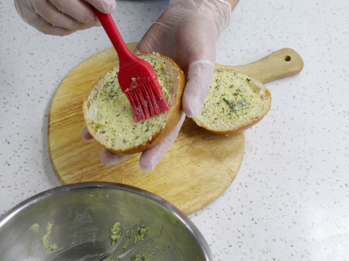 Una mano con guante usando un pincel de silicona rojo para esparcir mantequilla de ajo uniformemente sobre la mitad de un panecillo de hamburguesa cortado en una tabla de madera.