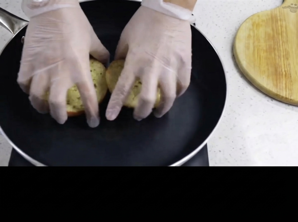 Manos con guantes colocando el lado con mantequilla de los panecillos de hamburguesa boca abajo en una sartén negra caliente.