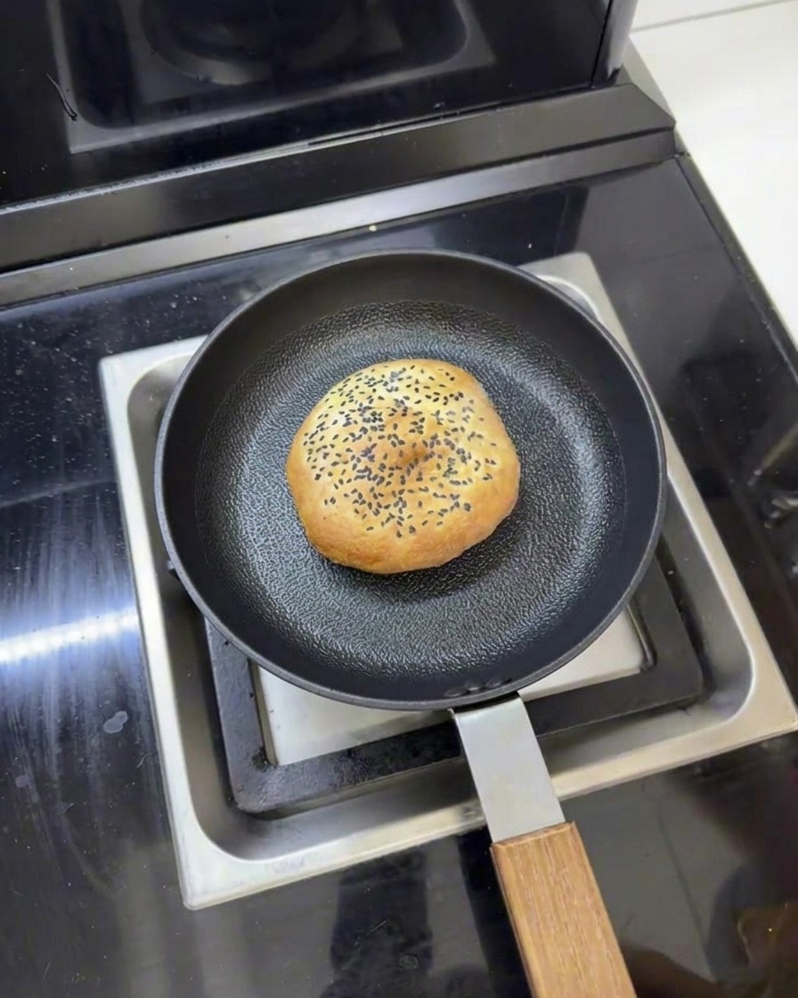 Un pan de hamburguesa de mantequilla en una sartén con agua alrededor, listo para ser calentado al vapor con tapa.