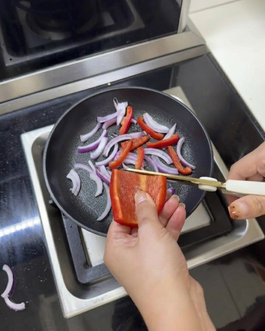 Cebollas rojas y pimientos en rodajas en una sartén siendo sazonados con sal y pimienta usando tijeras de cocina.
