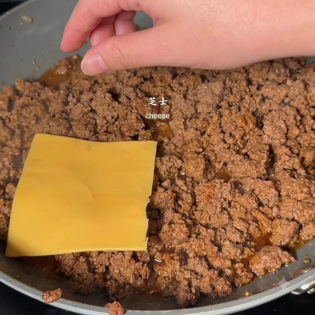 Una rebanada de queso amarillo colocada sobre la carne picada cocinada en una sartén por una mano.