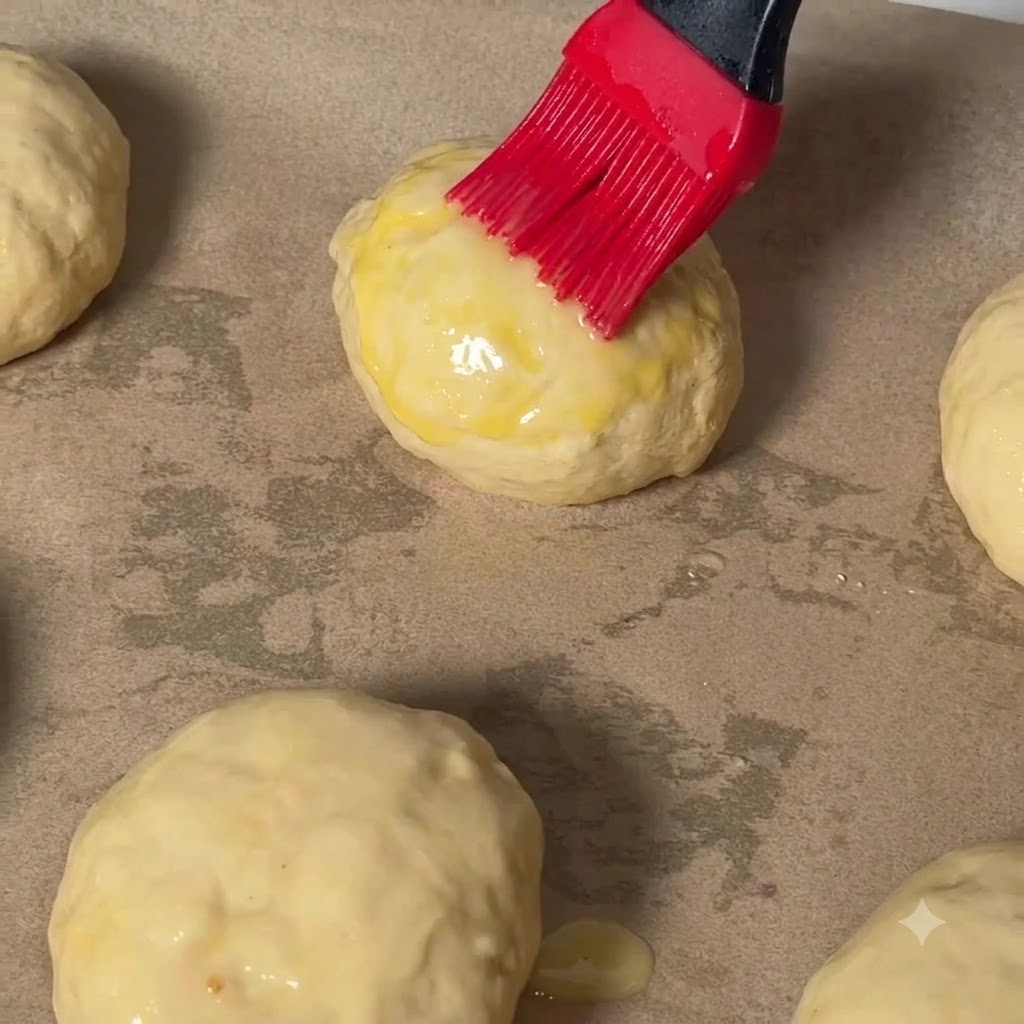 Una brocha de repostería roja aplicando baño de huevo en la parte superior de una bola de masa de hamburguesa sobre papel de horno.