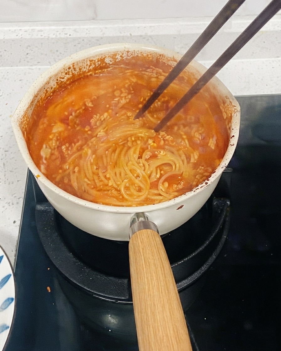 Una olla de pasta a base de tomate cociéndose a fuego lento en una estufa con palillos removiendo los fideos para espesar la salsa.