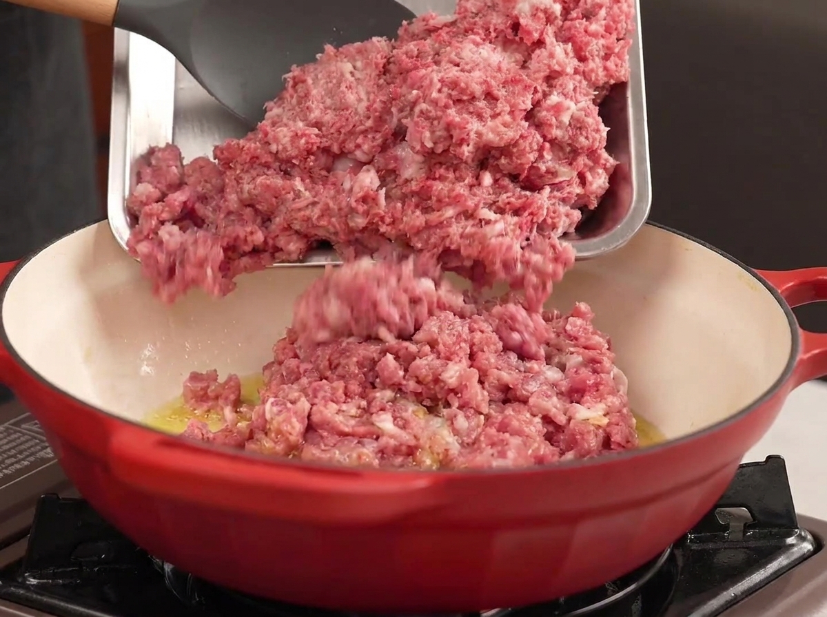 Carne picada de res y cerdo siendo vertida desde una bandeja de metal en una olla roja con aceite caliente.