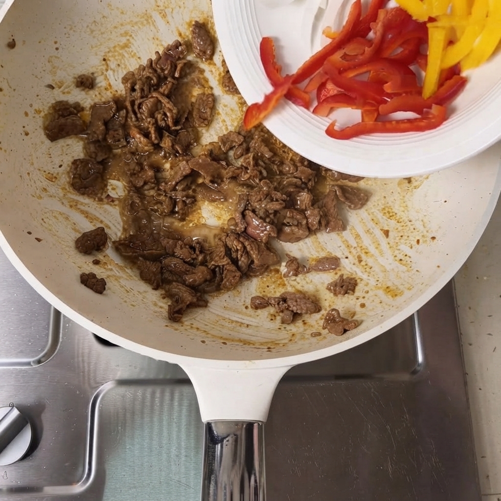Pimientos morrones rojos y amarillos en rodajas vertiéndose desde un plato blanco a una sartén con rodajas de ternera cocinadas.