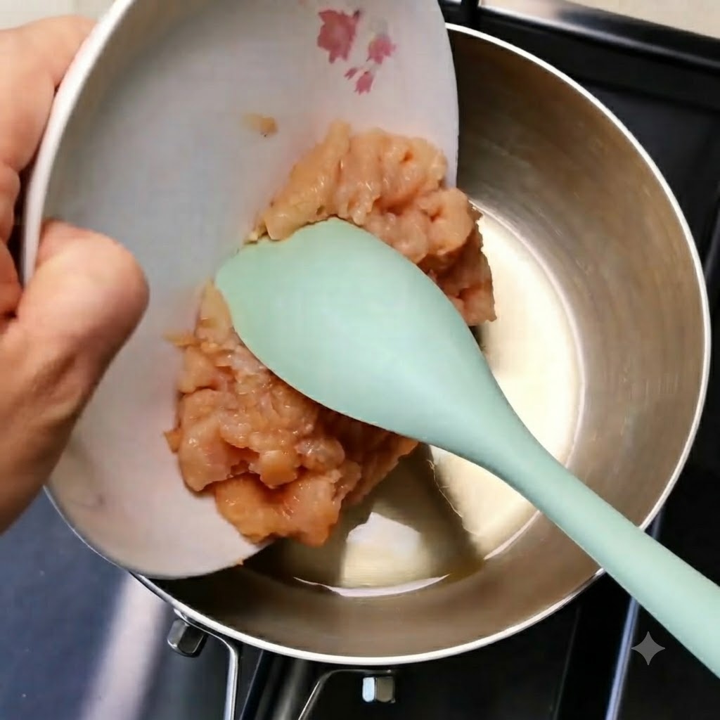 Carne de pollo picada deslizándose desde un plato blanco a una sartén metálica usando una espátula verde.