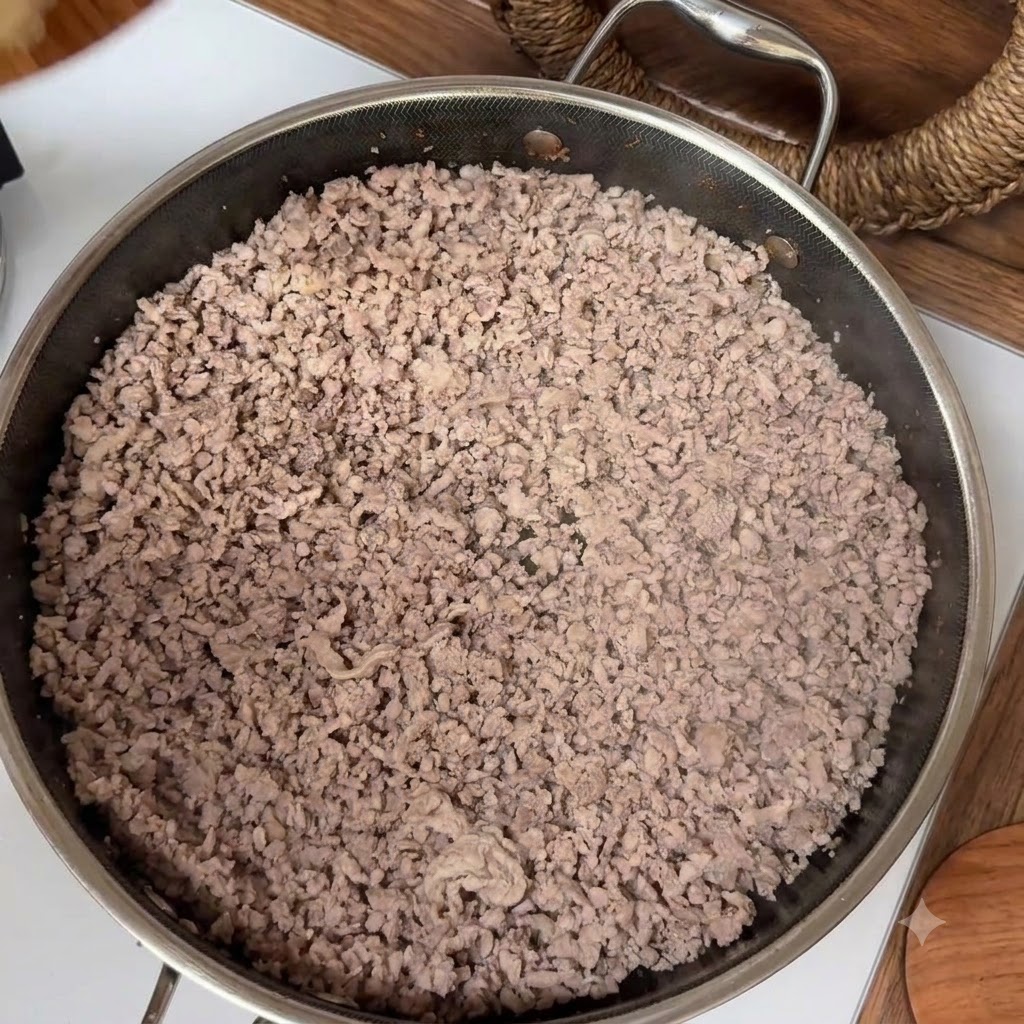 Carne picada cocinada llenando una sartén de acero inoxidable durante el proceso de sofrito.