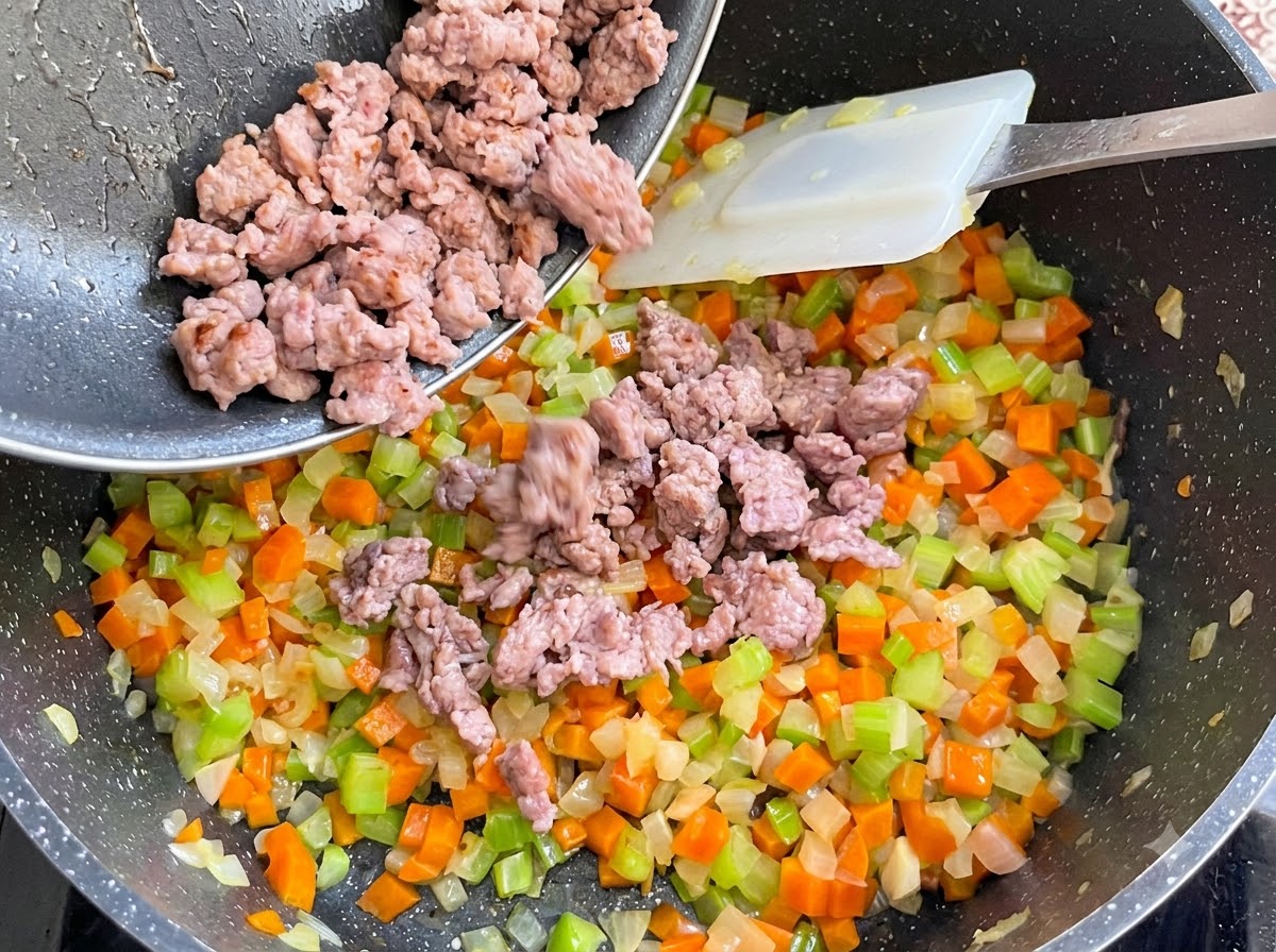 Carne de salchicha cocida siendo vertida desde una sartén a la olla de verduras y mezclada.